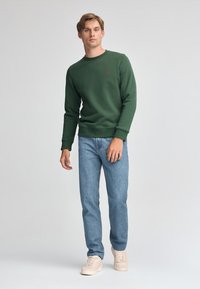 Grön crewneck sweatshirt med ribbat krage och ärmslut, tillsammans med ljusblå jeans. Modell med ljusbeiga sneakers, står mot en neutral bakgrund.