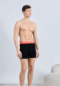 Άνδρας χωρίς πουκάμισο με κοντά σγουρά μαλλιά, φορώντας μαύρα boxer με κόκκινη μέση Calvin Klein, στέκεται δίπλα σε μια ανάγλυφη λευκή καρέκλα.