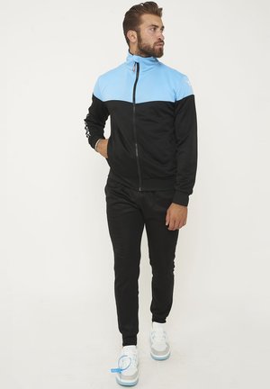 Uomo con giacca nera e blu con zip, pantaloni neri e sneakers bianche, in piedi con una mano in tasca, guarda verso destra.