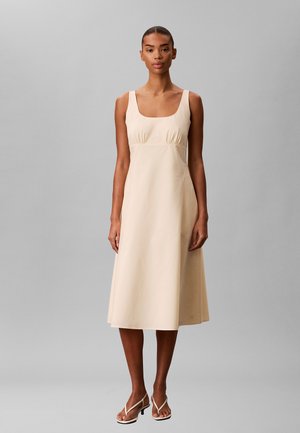 Vrouw staat frontaal, draagt een mouwloze beige midi-jurk en witte sandalen met hak en bandjes tegen een effen grijze achtergrond.