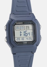 Digital Casio watch with a navy blue rubber strap, square black face, white LCD display, and silver buttons. Water-resistant up to 100 meters.