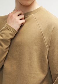 Sweat-shirt marron texturé avec un col rond et des manches raglan. Le tissu semble doux et épais, avec des détails de couture visibles.