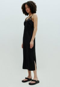 Zwart satijnen slipdress met dunne bandjes, kantdetails bij de halslijn en zijsplitten. Model draagt chunky zwarte sandalen.
