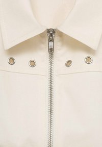 Giacca color crema con colletto a punta, zip metallica centrale e orecchini argento su entrambi i lati della zip. Tessuto dalla texture liscia.