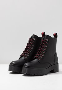 Botas de tobillo de cuero negro con cordones rojos, punta redonda, tacón alto y cuadrado, suela texturizada y tirador en la parte posterior. Presenta ojales de metal.