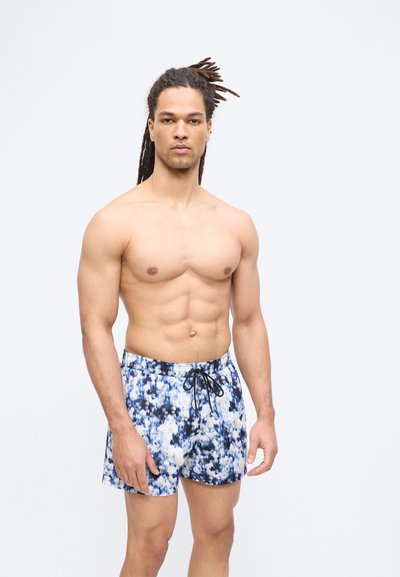 Άνδρας με ξυλάκια στα μαλλιά που φορά μπλε και λευκά μαγιό με σχέδια, στέκεται μπροστά σε ένα απλό φωτεινό φόντο.