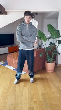 Casaco de capuz cinza com fecho, camisa branca, calças de ganga azuis escuras largas com punhos estampados, e tênis prateados. Ambiente interior com uma planta e móveis.