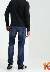 Svart sweatshirt, blå denimjeans med något avsmalnande passform och svarta sneakers med vita detaljer och en logotyp. Enkelt bakgrund.