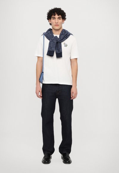 Balts T-krekls ar zaļu logo, jūras zila rakstaina džemperis, kas sasieta ap pleciem, tumši zili džinsi un melni apavi. Ikdienišķs apģērbs ar aksesuāriem.