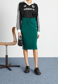 Musta urheilupaita valkoisella kauluksella ja kolmella raidalla, yhdistettynä vihreään neulottuun kynähameeseen, mustiin loafereihin ja pieneen mustaan käsilaukkuun.