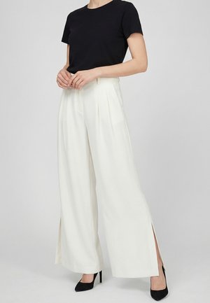 Persona que lleva una camiseta negra de manga corta, pantalones blancos de pierna ancha y talle alto con aberturas laterales, y tacones negros de punta puntiaguda, de pie sobre un fondo gris.