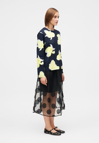 Μπλε τοπ με μακριά μανίκια και κίτρινο λουλουδάτο μοτί�βο, συνδυασμένο με διαφανή μαύρη φούστα με μεγάλα πουά και μαύρες φλατ μπότες με διακοσμητικά στοιχεία.