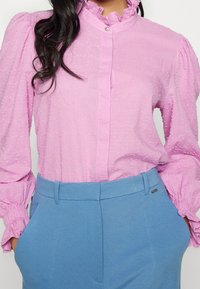 Pink blouse with textured dots, ruffled collar, and puffed sleeves paired with light blue high-waisted trousers featuring side pockets.