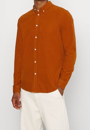 Mężczyzna ubrany w pomarańczową koszulę z długim rękawem zapinaną na guziki oraz białe spodnie, stojący z jedną ręką w kieszeni na jednolitym tle.