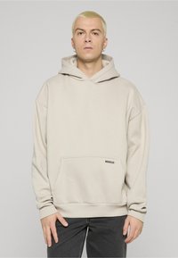 Gaiši bēša kapuce ar kenguras kabatu, ribota apmales aprocēm un piešūtu kapuci. Priekšējā apakšmalā ir neliela melna logotipa etiķete.