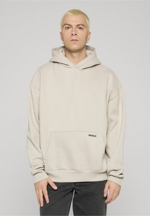 Ljusbeige hoodie med kenguruficka, ribbade muddar och fastsydd huva. Har en liten svart logotypetikett på framkanten.