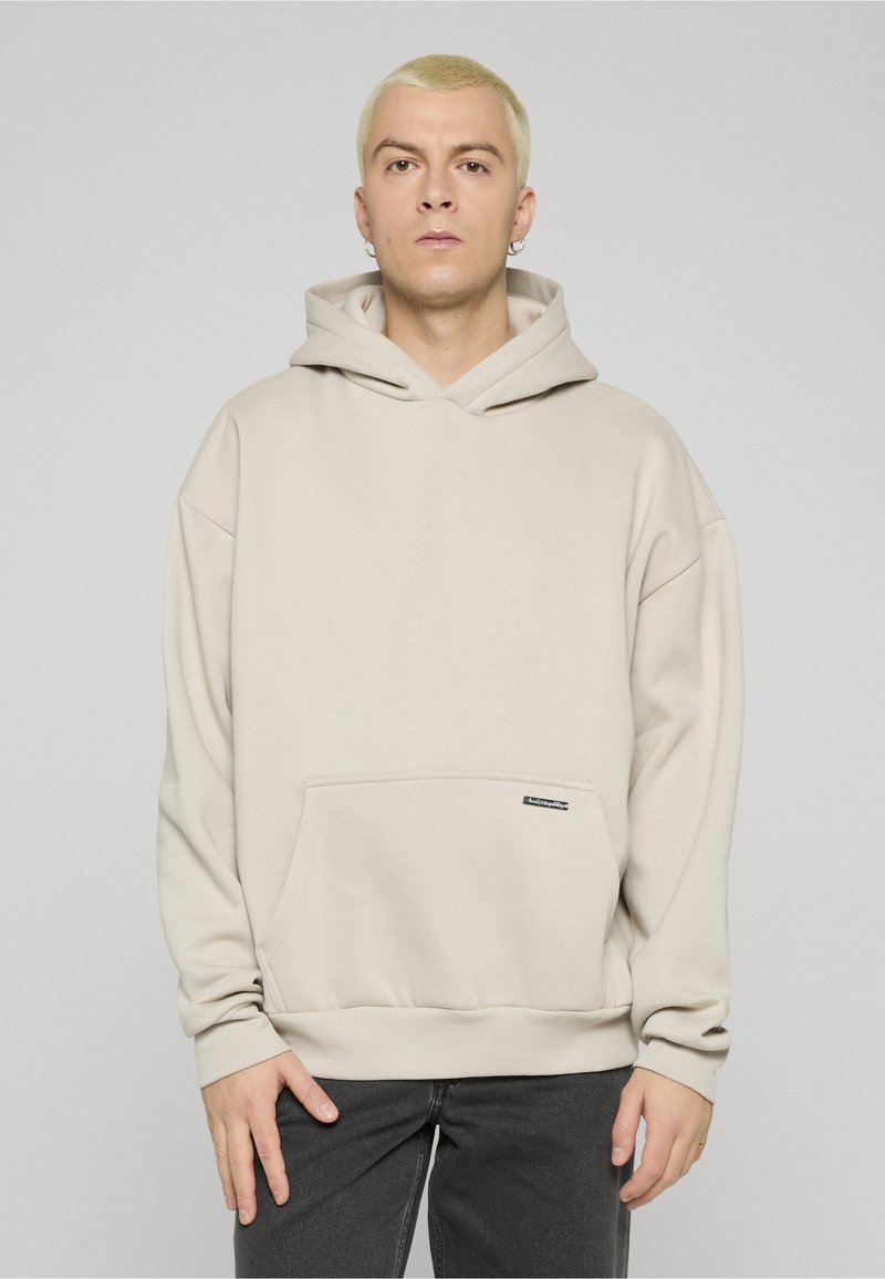 Gaiši bēša kapuce ar kenguras kabatu, ribota apmales aprocēm un piešūtu kapuci. Priekšējā apakšmalā ir neliela melna logotipa etiķete.