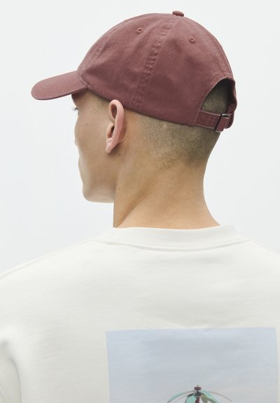 Man draagt een vervaagde rode honkbalpet en een witte sweatshirt met een afbeelding van een watertoren en gebouwen op de achterkant, kijkt weg.