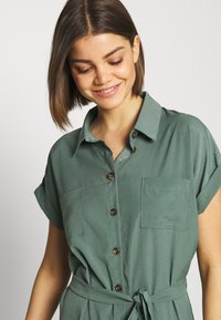 Vestito camicia verde con bottoni e maniche corte, due tasche sul petto e vita aderente. Presenta bottoni marroni e una texture del tessuto liscia.