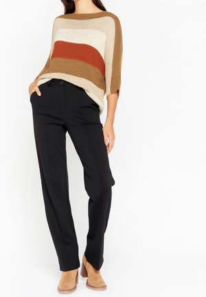 Mujer con un suéter de punto suelto con rayas beige, blancas, color óxido y marrones, pantalones negros y botines de ante color beige con tacón grueso.