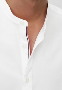 Vit skjorta med mandarin-krock, med en enda knapp och kontrasterande röd och svart söm längs knappslået. Slät textur.