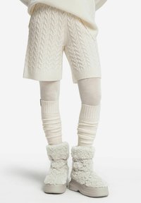 Białe szorty dzianinowe w warkocze i pasujące ocieplacze na nogi, w zestawieniu z puszystymi białymi botkami na grubej podeszwie. Tekstylne i miękkie materiały.