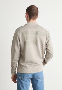 Beige genser med ribbestrikkede mansjetter og kant, med et teksturert logo i lysegrå på baksiden. Et par blå jeans synlige.