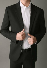Blazer nero con un solo bottone, bavero cucito, indossato su una camicia bianca con colletto. Pantaloni scuri e cintura completano l'outfit.