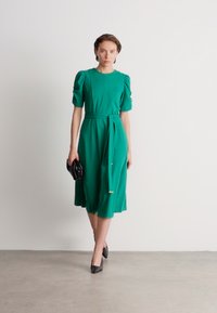 Πράσινο φόρεμα με κοντά φουσκωτά μανίκια, ζωνάκι στη μέση και μήκος μίντι. Λείο ύφασμα με ελαφριά λάμψη; συνδυάζεται με μαύρη τσάντα και τακούνια.