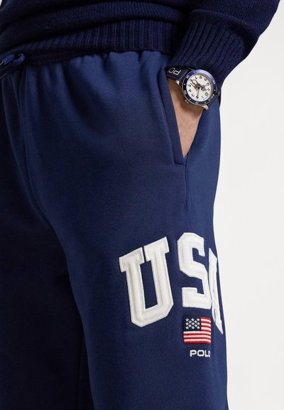 Marineblaue Jogginghose mit weißem "USA"-Schriftzug und einem Aufnäher der amerikanischen Flagge. Strukturierter Stoff, ausgestattet mit einem Kordelzugbund und Seitentaschen.