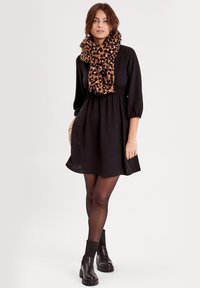 Robe noire avec des manches trois-quarts et une taille élastique, associée à une écharpe imprimée léopard. Le mannequin porte des bottines noires et des collants.