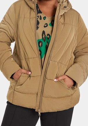 Personne portant une veste matelassée beige avec les mains dans les poches, dévoilant un top à motif léopard marron et vert en dessous.
