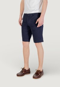 Pantaloni corti in cotone blu navy con design a frontale piatto, dotati di tasche laterali e orlo dritto. Abbinati a scarpe da barca in pelle marrone.