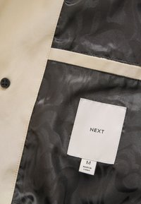 Interior de una chaqueta de tela beige con forro negro estampado. Presenta una etiqueta que dice "NEXT," talla "M," y una etiqueta con información de cuidado. Botón negro visible.