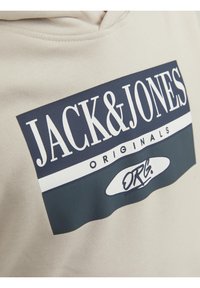 Sweat à capuche beige avec un logo rectangulaire noir et blanc indiquant « JACK & JONES ORIGINALS ORG. » sur la poitrine.