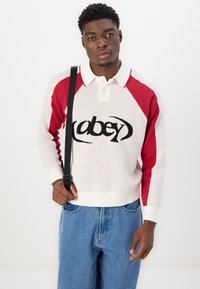 Röd och vit långärmad polo med kontrasterande ärmar, som har en stor svart "obey"-logotyp, texturerad stickad tyg och en knäppt krage.