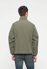 Chaqueta bomber verde con cuello alto y puños elásticos, con una textura suave y un corte relajado. Combinada con jeans de mezclilla oscura.