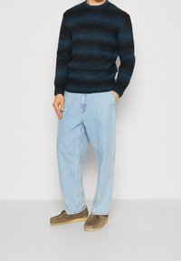 Maglione a righe blu, jeans in denim blu chiaro e scarpe in suede marroni. Il maglione presenta una trama a coste e un colletto a girocollo.