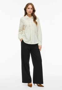 Vrouw met lang bruin haar draagt een crèmekleurige gestreepte blouse met knopen en losse zwarte broeken met bruine puntige hakken, staand met één hand in de zak.