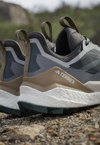 Close-up van grijze en bruine Terrex trailrunning schoenen die rusten op rotsachtige grond buiten.