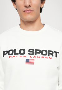 Felpa bianca con la scritta "POLO SPORT" in grassetto nero, "RALPH LAUREN" in rosso, e un piccolo dettaglio della bandiera americana sotto il testo.