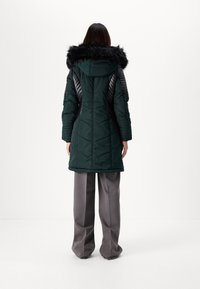 Long green puffer coat with a fur-lined hood, quilted texture, and contrasting black shoulder panels. Paired with gray wide-leg trousers.