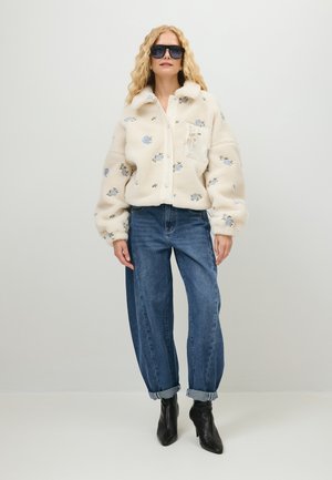 Vrouw draagt een oversized crèmekleurig fleecejack met blauwe bloemenprints, opgerolde blauwe spijkerbroek, zwarte laarzen met hak en grote zwarte zonnebril.