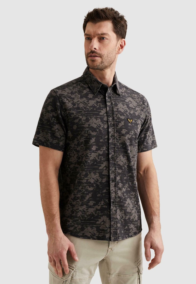 Homme portant une chemise à manches courtes noire et grise à motif camouflage avec un petit logo jaune sur la poitrine, pantalon cargo beige.