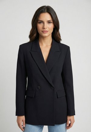 Vrouw met donker haar, gekleed in een zwarte dubbelgeknoopte blazer en lichtblauwe spijkerbroek, staand tegen een eenvoudige lichte achtergrond.