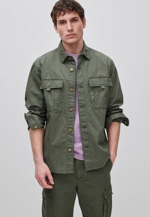 Młody mężczyzna stoi zwrócony przodem, ubrany w oliwkowo-zieloną roboczą kurtkę zapinaną na guziki, na jasnofioletową koszulę oraz pasujące oliwkowo-zielone spodnie typu cargo.