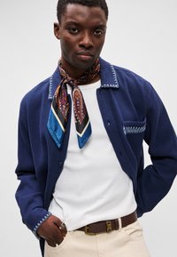 Ναυτικό πουκάμισο με υφή και αντίθετο μπλε γιακά, λευκό T-shirt από κάτω, καφέ ζώνη και μοτίβο μεταξωτό μαντήλι δεμένο γύρω από τον λαιμό.