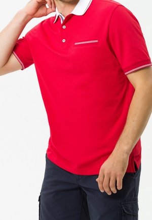 Uomo che indossa una polo rossa a maniche corte con colletto bianco e pantaloncini cargo blu navy, posando con una mano vicino al collo e l'altra rilassata.