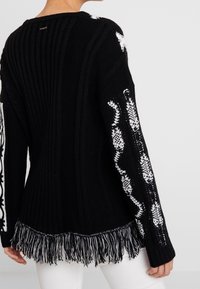 Black knitted sweater with white patterned accents on the sleeves, featuring ribbed texture and a fringed hemline.