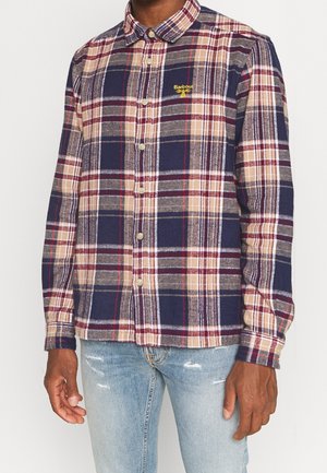 Mężczyzna w niebieskiej, beżowej i czerwonej kraciastej koszuli z żółtym logo Barbour oraz jasnoniebieskich, postarzanych dżinsach na białym tle.